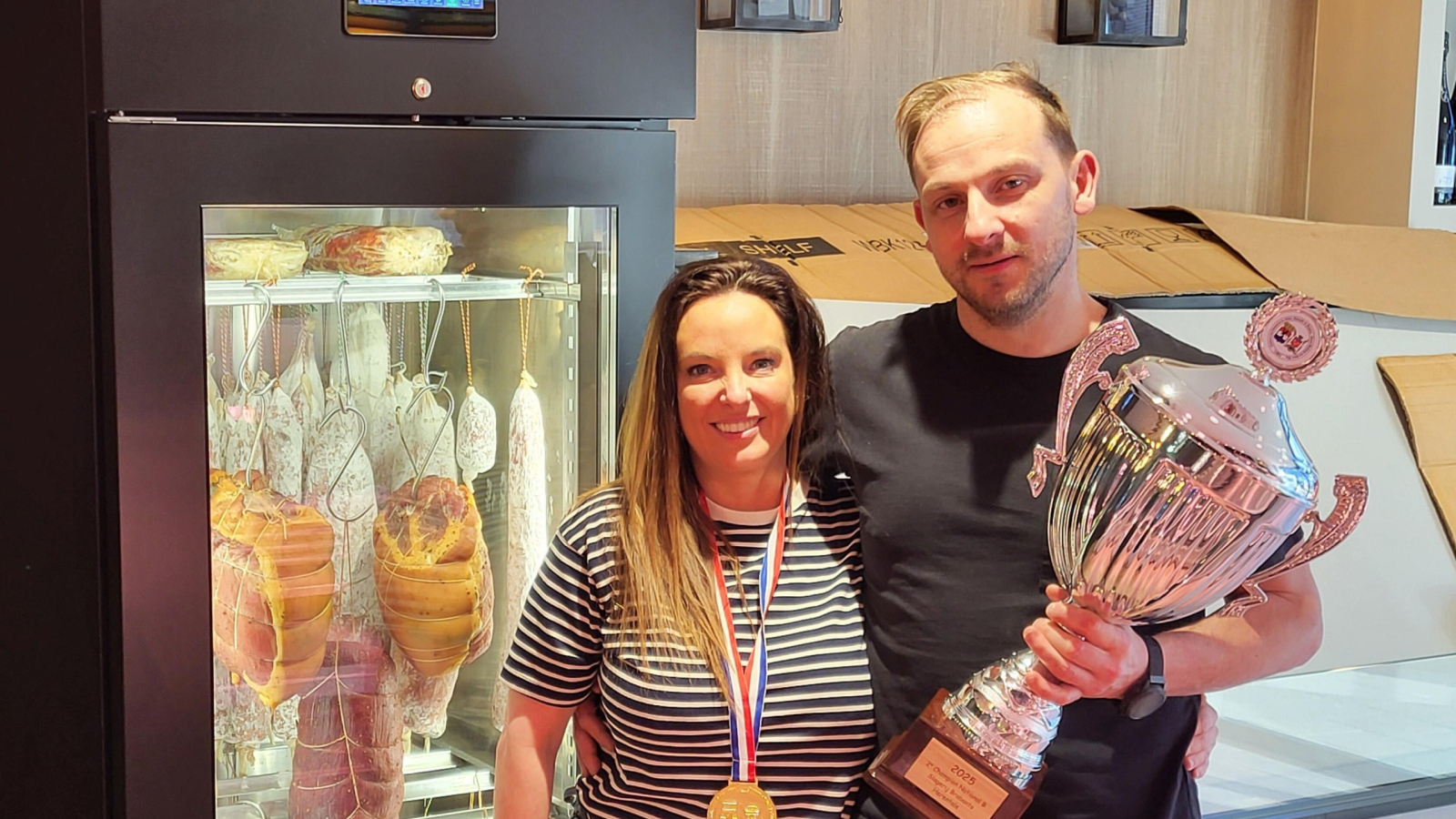 Jelle en Evy Brabants van culinaire slagerij Brabants (Herentals) poseren met trofee en medaille voor hun nieuwe klimaatkast vol vleeswaren.