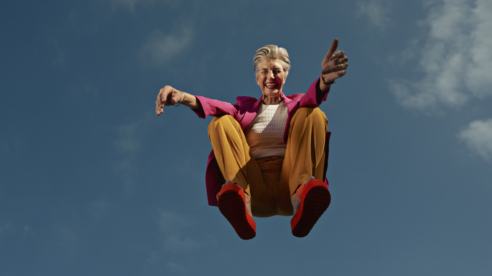 Happy senior woman crouching mid-air against blue sky