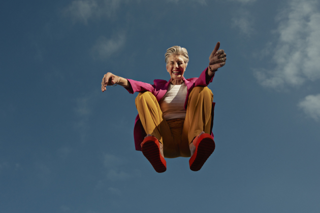 Happy senior woman crouching mid-air against blue sky