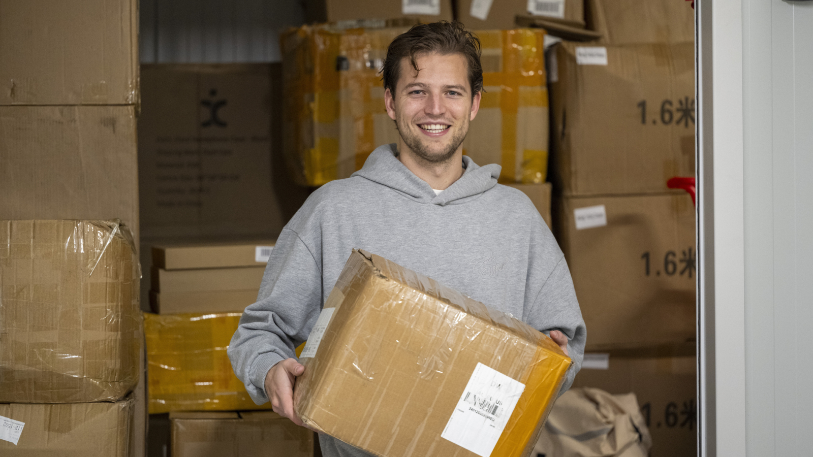 Smiling man in a grey hoodie holds a cardboard box in a storage area filled with stacked boxes, Molenhoek, Netherlands