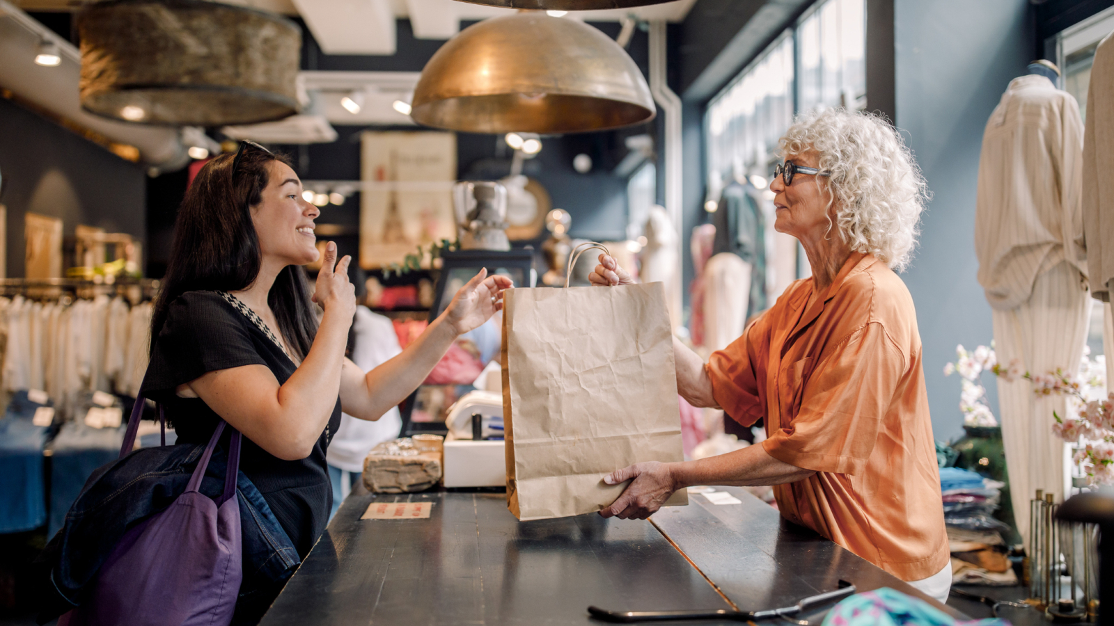 Senior female owner giving bag to customer at checkout in clothing store