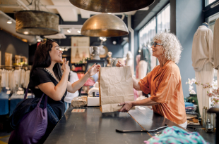 Senior female owner giving bag to customer at checkout in clothing store