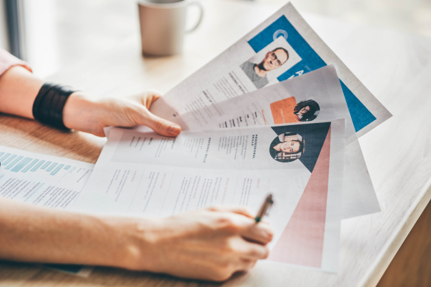 A recruiter’s hands hold a fan of printed resumes over a wooden desk. This shot represents human resources, job hunting, and the professional hiring process.