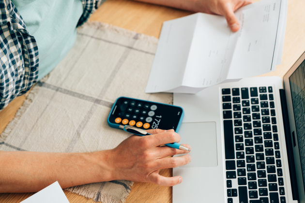 Unrecognizable person counting expenses on calculator in mobile phone app, checking her energy bills at home. Budget management, price increases, inflation, rise in price of products and utilities. View from above, managing home finances