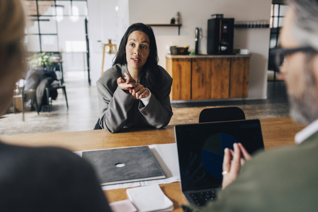 Woman gesturing while giving interview to business experts while sitting at table in office